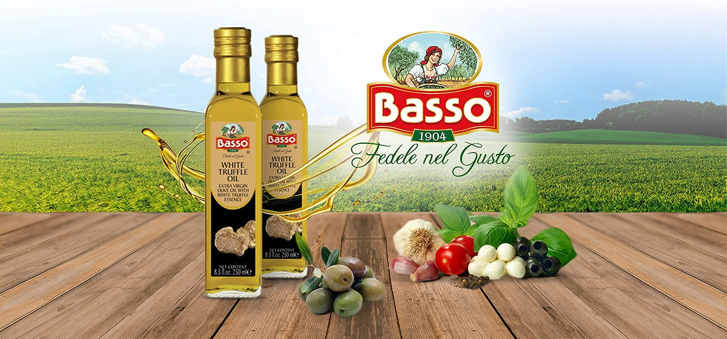 Two bottles of Basso White Truffle Extra Virgin Olive Oil displayed on a wooden surface with fresh ingredients like olives, garlic, basil, tomato, and cheese, set against a scenic green field background with the Basso logo and slogan "Fedele nel Gusto".