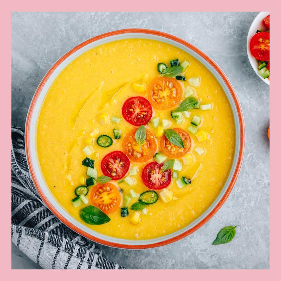 Bowl of creamy yellow tomato gazpacho garnished with fresh red and yellow cherry tomato slices, diced cucumber, corn, basil leaves, and green chili on a gray table with a striped napkin.