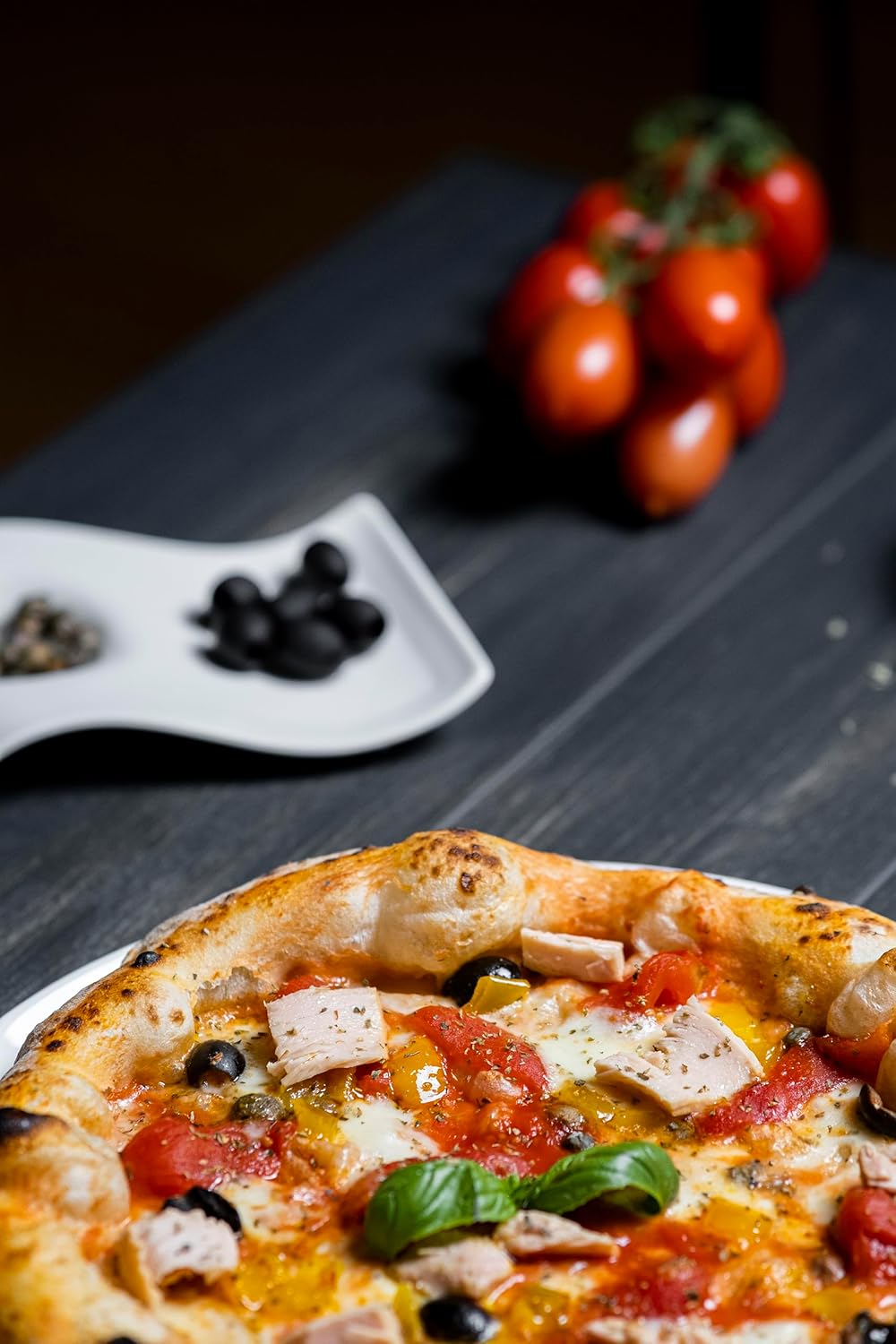 Close-up of an Italian pizza topped with tuna, mozzarella, olives, and fresh basil, with vine-ripened red Datterino tomatoes and a dish of black olives in the background.
