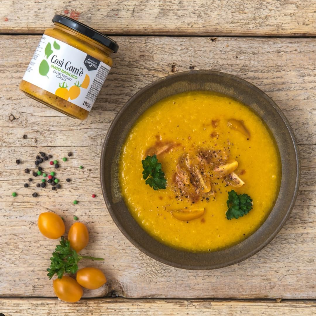 Bowl of yellow tomato soup garnished with parsley and pepper, next to a jar of Così Com’è Yellow Datterino Tomato Sauce with Basil on a rustic wooden table, surrounded by fresh yellow tomatoes and peppercorns.