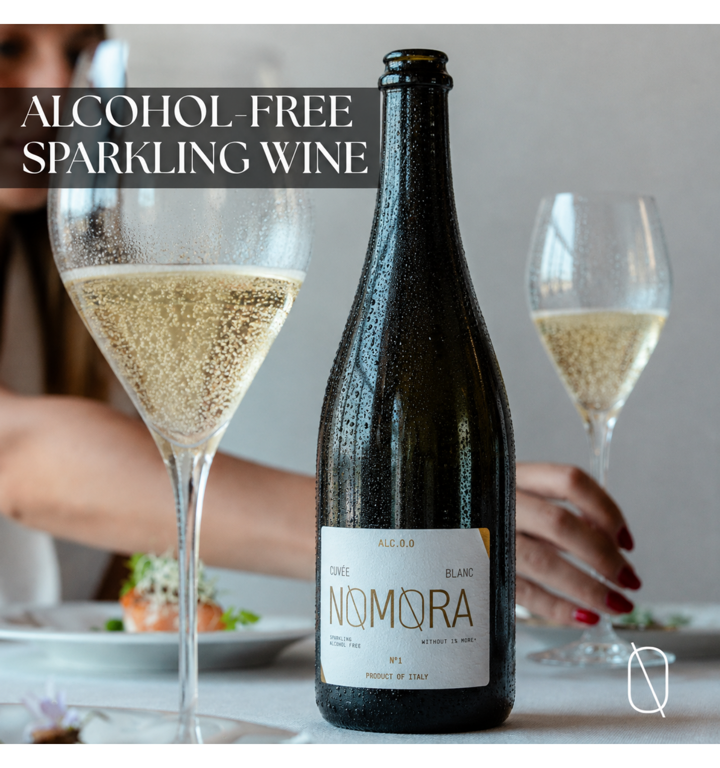 A bottle of Nomora alcohol-free sparkling wine stands on a table between two filled wine glasses, with a person's hands and blurred figure in the background. Text at the top reads 'ALCOHOL-FREE SPARKLING WINE'.