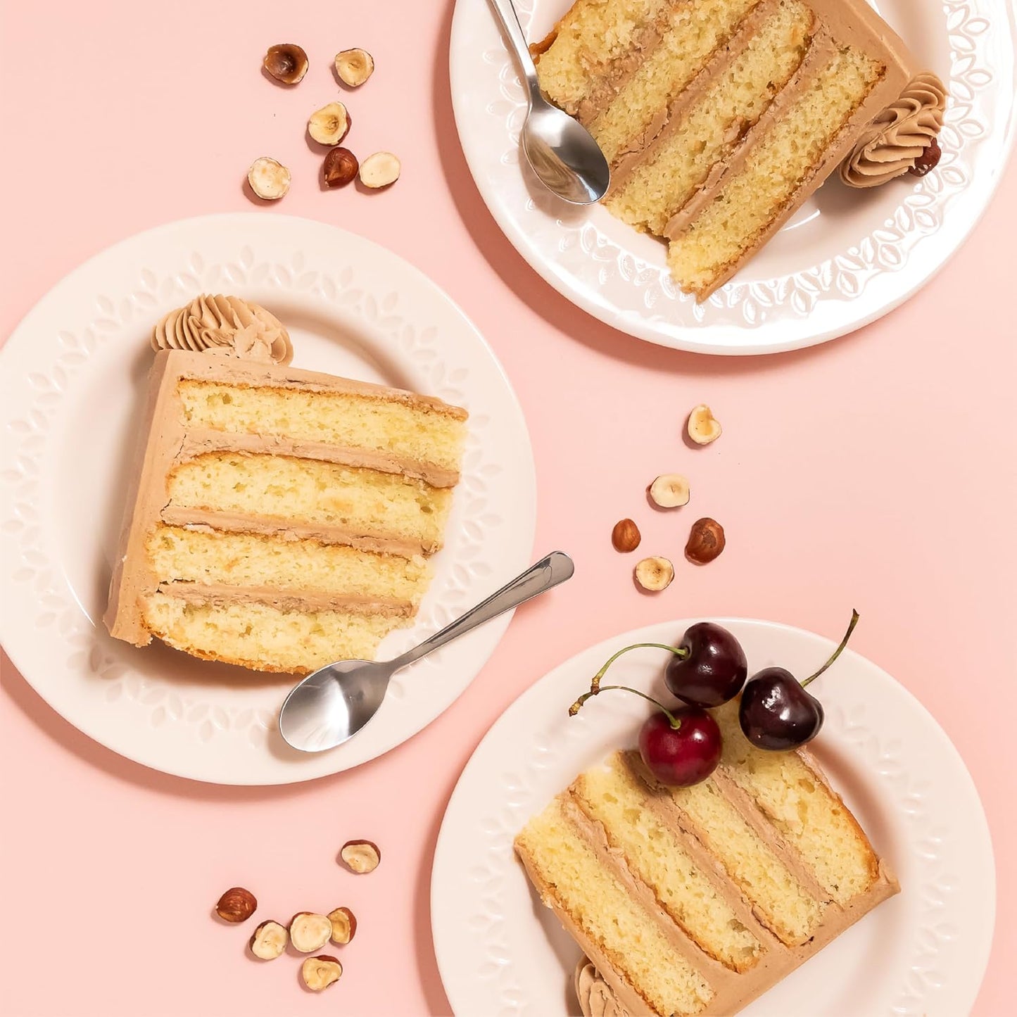 Plates with slices of layered vanilla cake filled with hazelnut praline cream, garnished with cherries and hazelnuts on a pink background.
