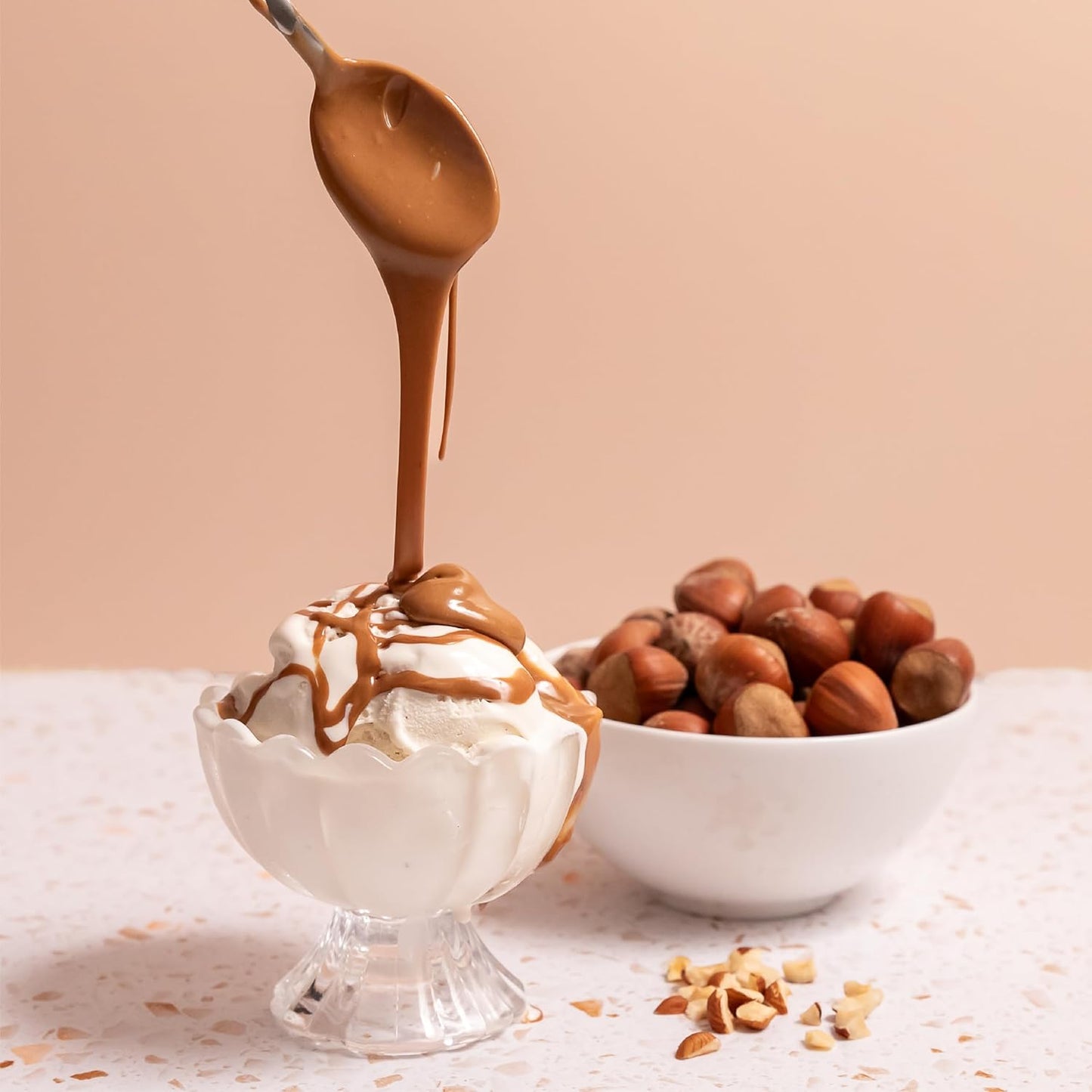 Hazelnut praline spread being drizzled over vanilla ice cream in a glass bowl, with a bowl of whole hazelnuts in the background.