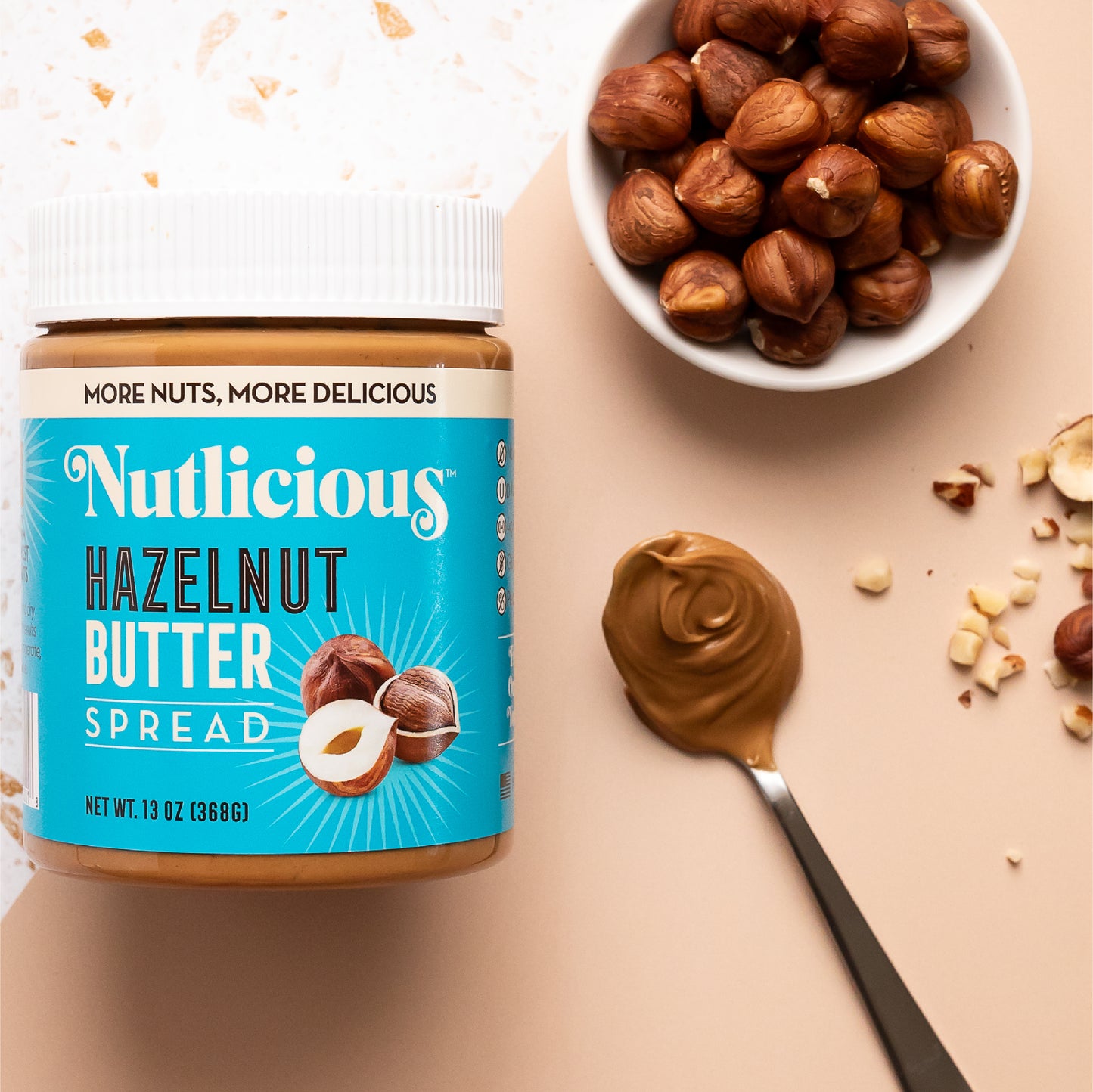 Nutlicious Hazelnut Butter Spread jar (13 oz) on a flat surface, styled with a bowl of roasted hazelnuts, a spoonful of creamy hazelnut butter, and scattered nut pieces.