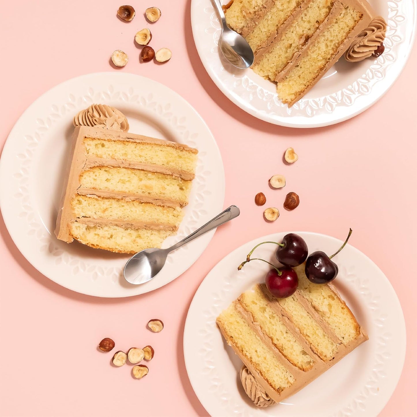Plates with slices of hazelnut-flavored layer cake filled and frosted with Nutlicious spread, decorated with cherries and hazelnuts on a pink background.