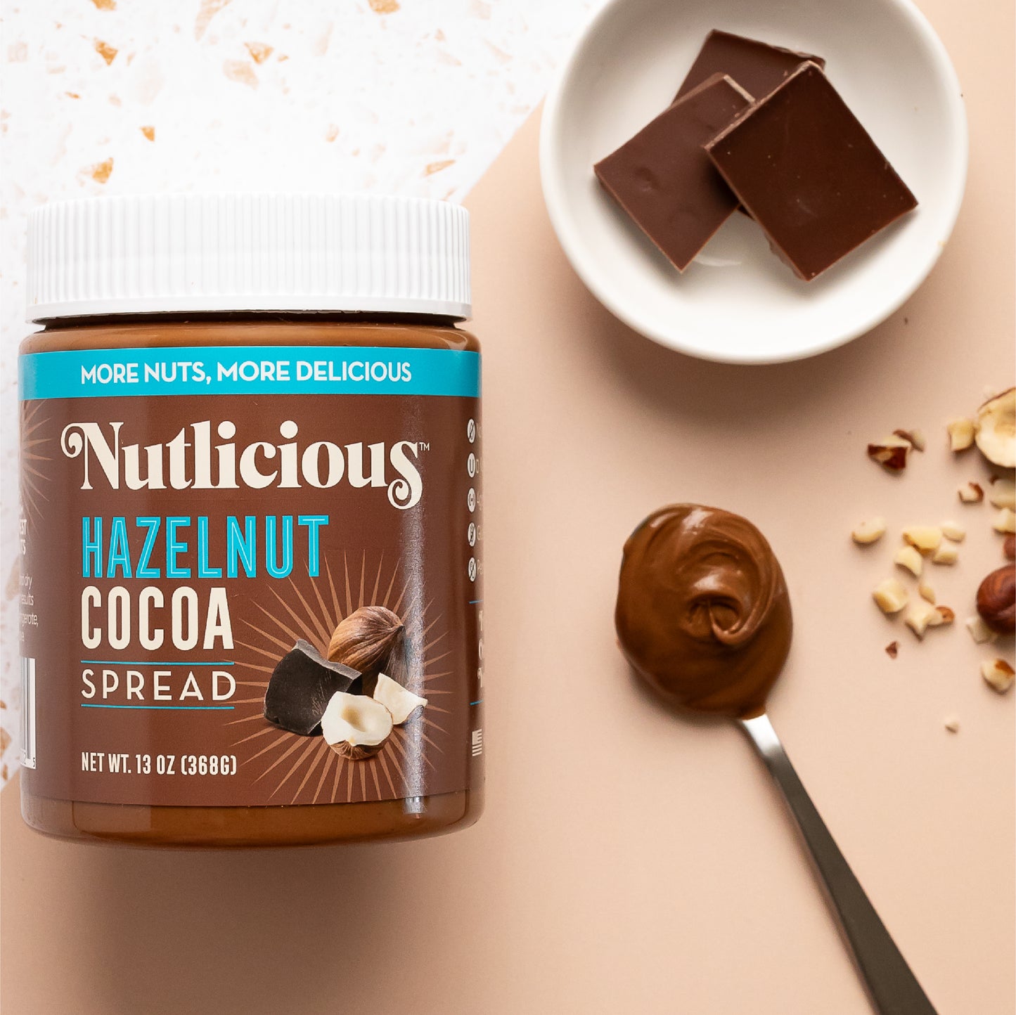 Nutlicious Hazelnut Cocoa Spread (13 oz) jar displayed on a flat surface with a spoonful of creamy spread, chocolate squares in a small dish, and scattered chopped hazelnuts for decoration.