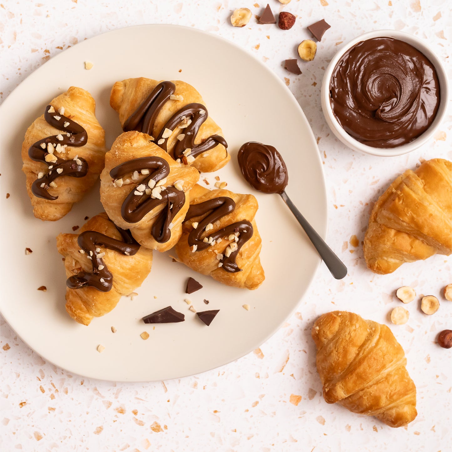 Freshly baked croissants on a plate drizzled with Nutlicious Hazelnut Cocoa Spread, topped with chopped hazelnuts, served with a spoonful of spread and a bowl of rich hazelnut cocoa cream.