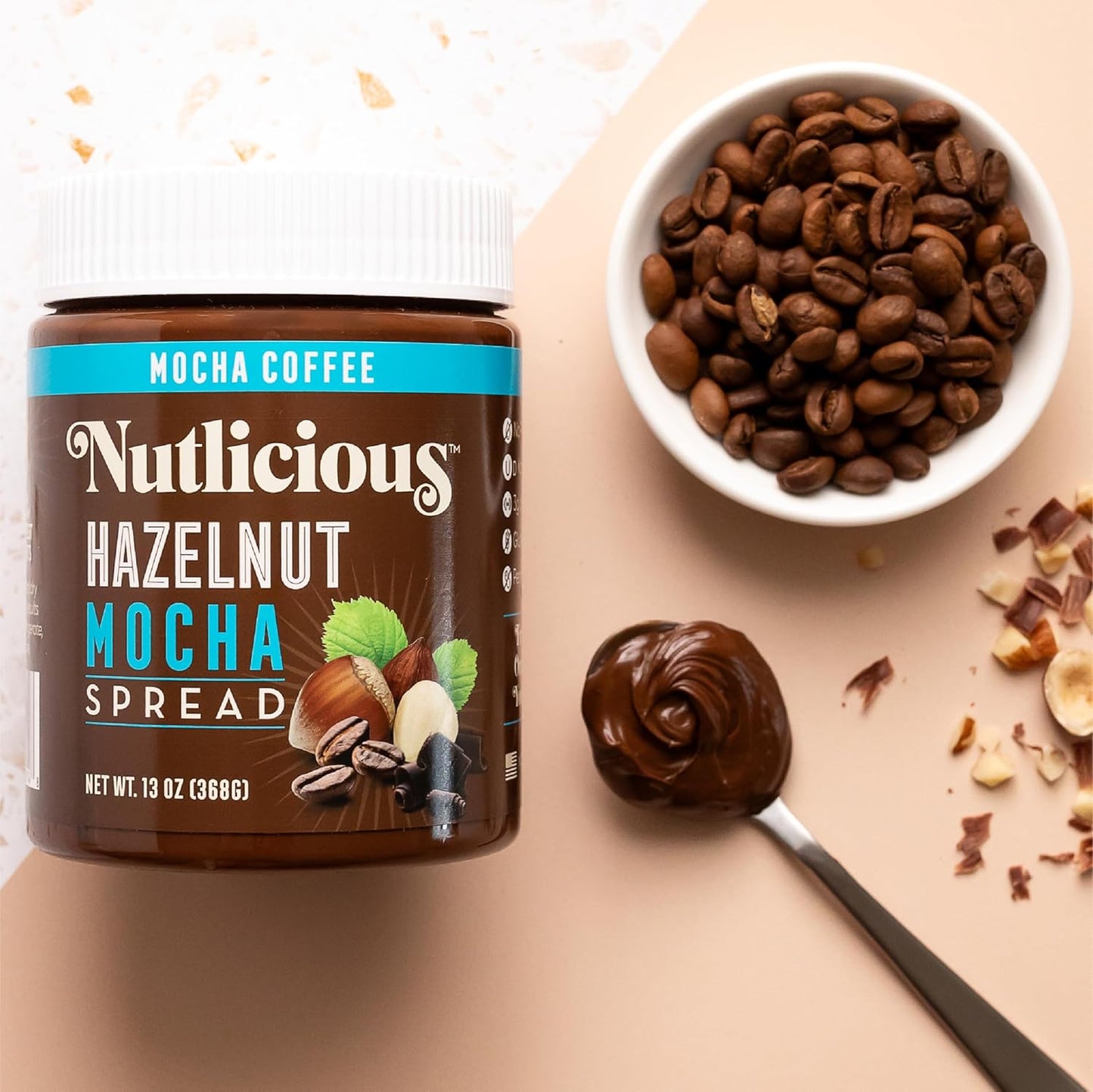 Nutlicious Hazelnut Mocha Spread jar with coffee beans in a bowl, a spoonful of creamy spread, and scattered hazelnut pieces on a beige background.