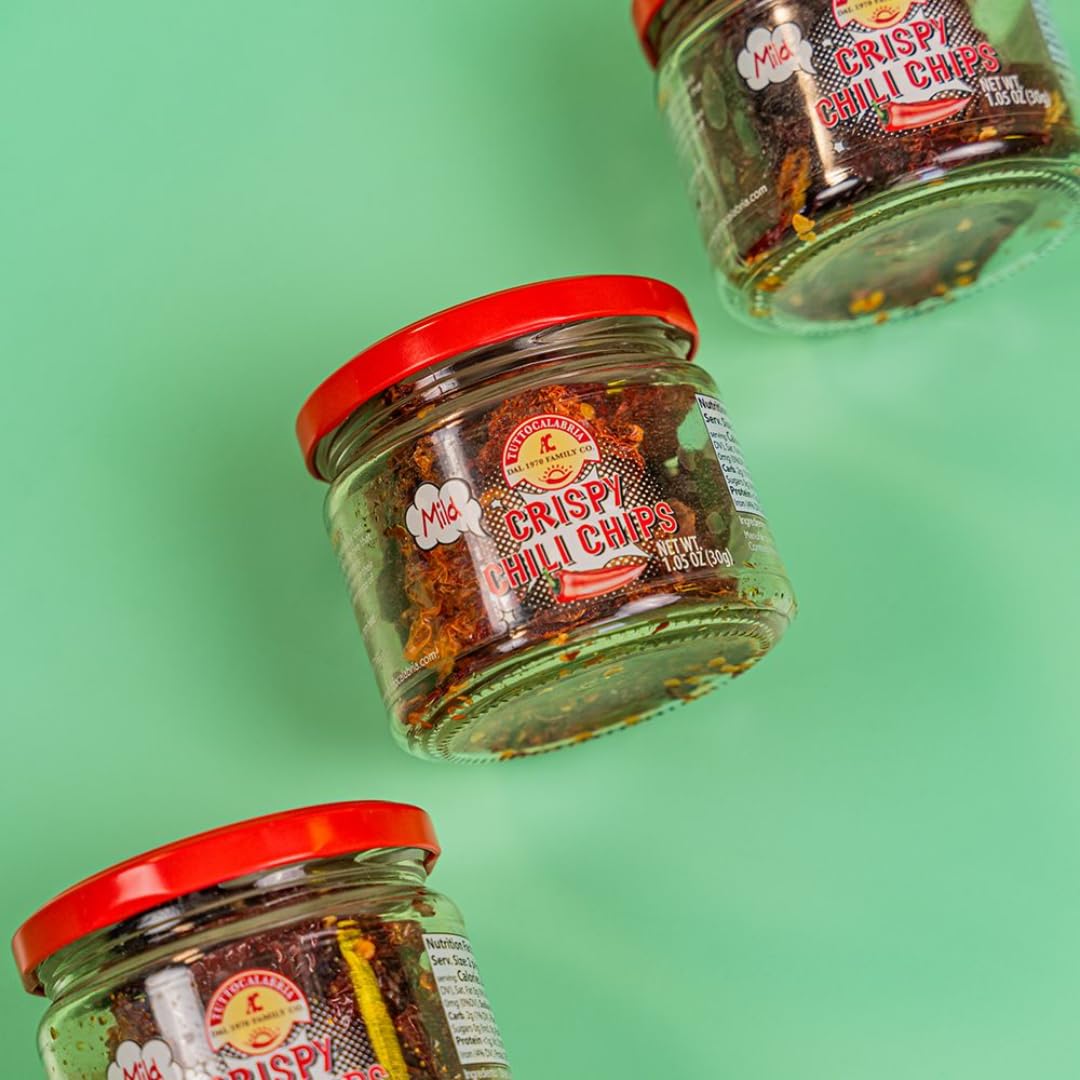 Three jars of TuttoCalabria Mild Crispy Chili Chips with red lids, displayed on a green background, arranged in a flat-lay style