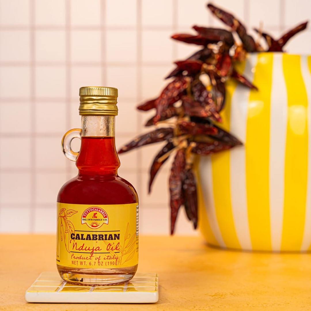 Bottle of Calabrian Nduja Oil on a coaster with a yellow and white striped container and dried herbs in the background.