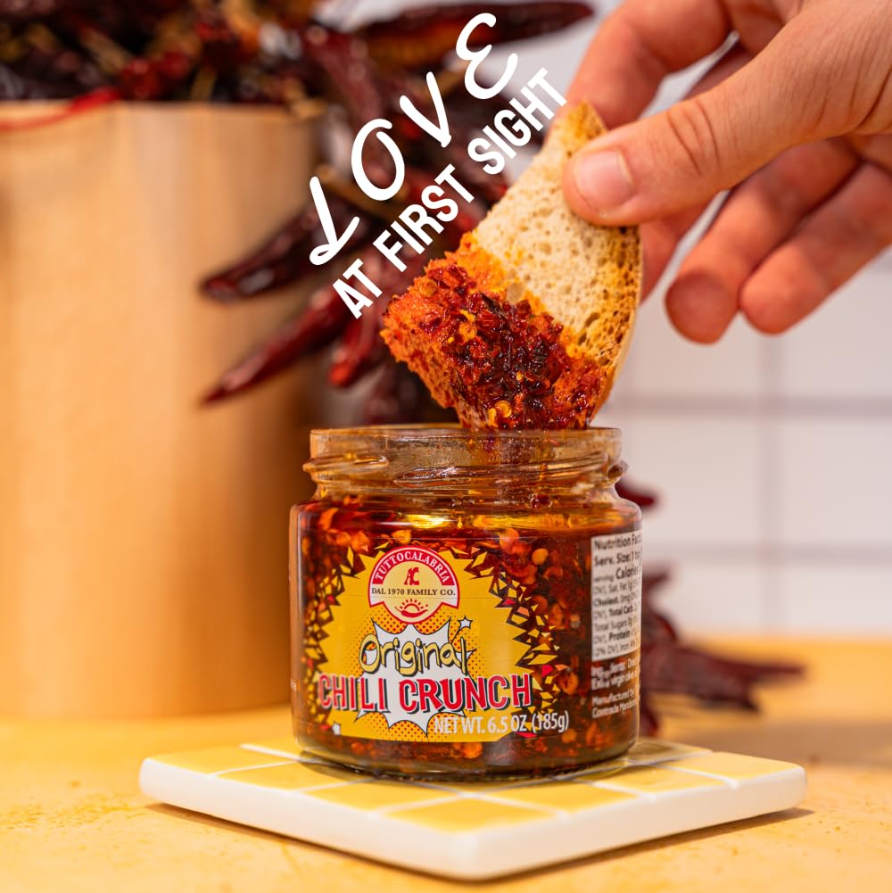 TuttoCalabria Hand dipping a cracker into a jar of chili crunch with text overlay on a wooden surface.