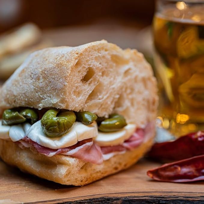 Rustic Italian sandwich with green olives, mozzarella cheese, and prosciutto on crusty bread, served on a wooden board with a drink in the background.