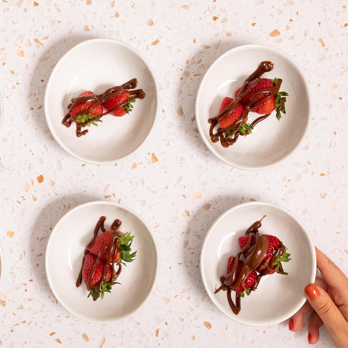 Four white plates with strawberries and chocolate sauce on a light speckled surface.
