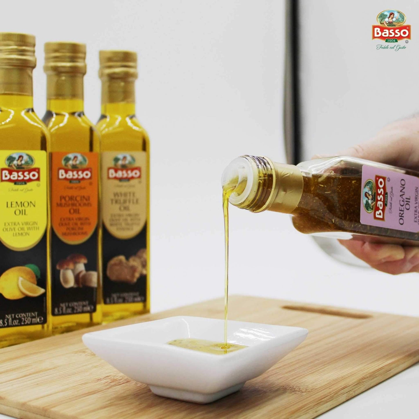 Hand pouring Basso 1904 Oregano Infused Extra Virgin Olive Oil into a white dipping bowl, with Lemon, Porcini Mushroom, and White Truffle flavored olive oil bottles displayed in the background.