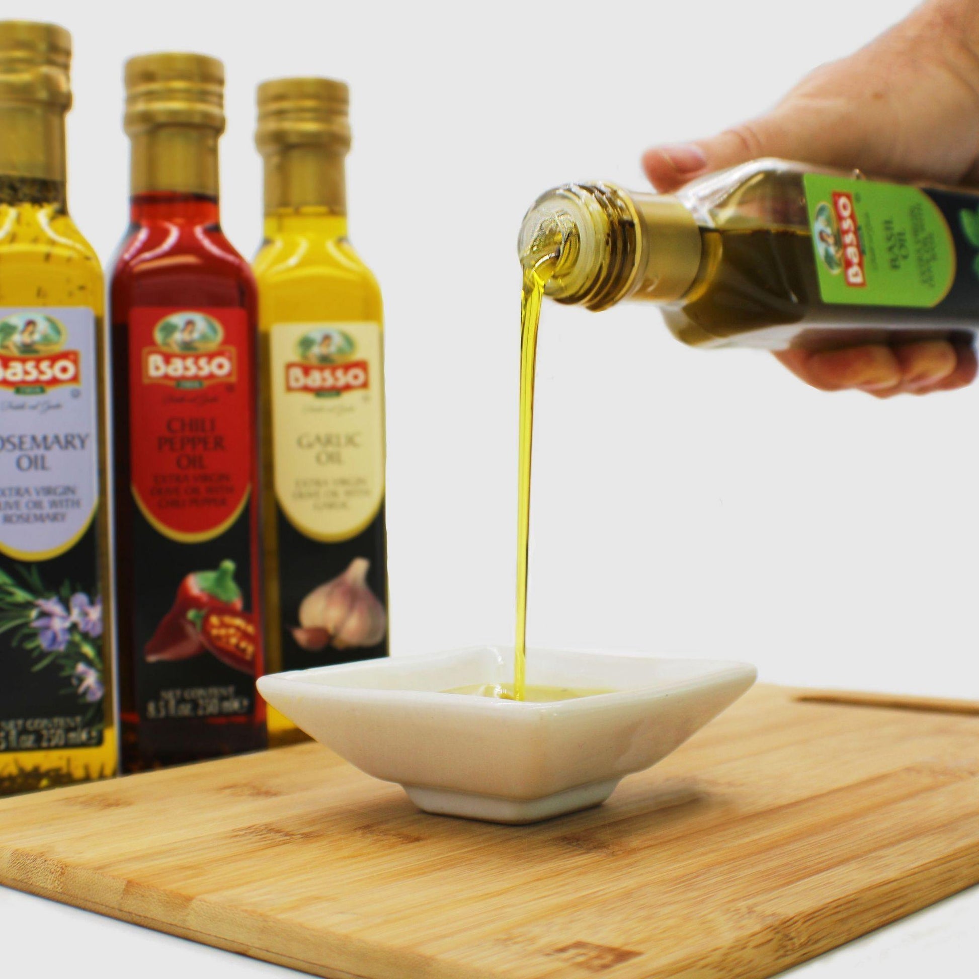 Hand pouring Basso flavored olive oil into a white dish with basil, rosemary, garlic, and chili infused olive oils in bottles.
