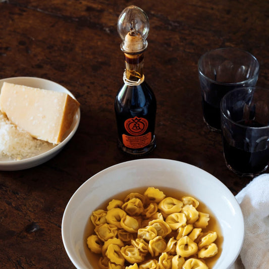 Bowl of tortellini with a bottle of red wine and a block of cheese on a wooden table.