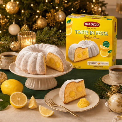 A festive holiday scene featuring a yellow lemon cake on a pedestal and a slice on a plate, alongside a yellow Balocco Torte in Festa box. The setting includes a decorated Christmas tree, lemons, cups of coffee, and gold ornaments on a dark green tablecloth.
