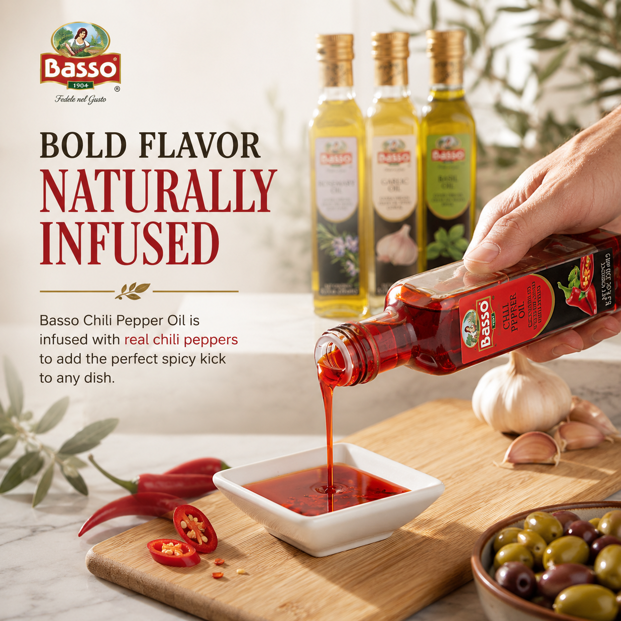A hand pours Basso Chili Pepper Oil from a glass bottle into a small white dish on a wooden cutting board, with Basso olive oil bottles, garlic, red chili peppers, and olives in the background and on a marble surface.