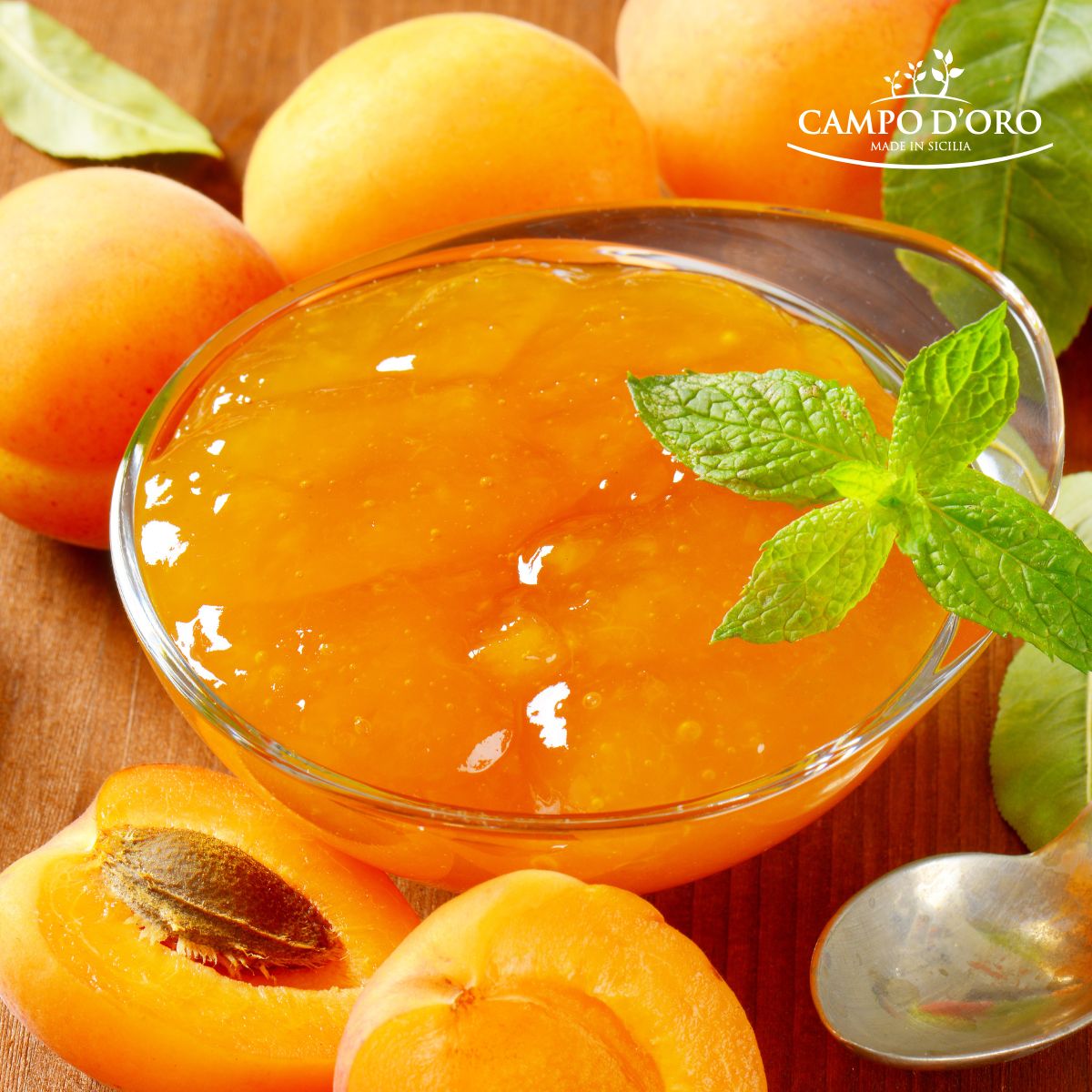Glass bowl of apricot jam with fresh apricots and mint leaves on a wooden surface, featuring the Campo d'Oro brand.