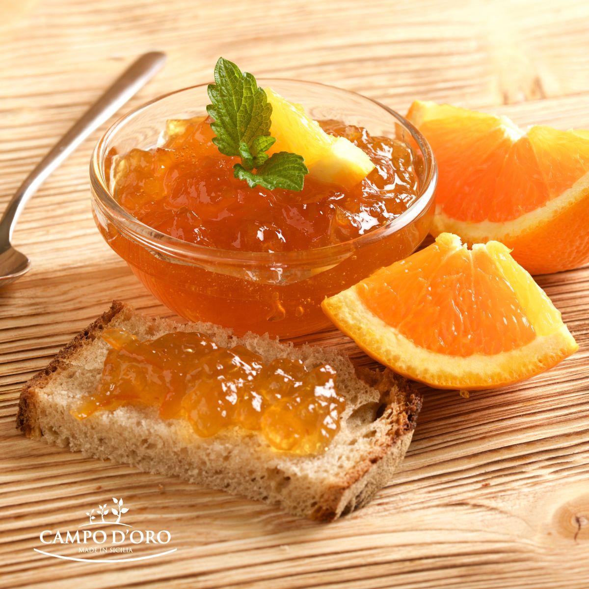 Glass bowl of orange marmalade with a slice of bread, orange slices, and a sprig of mint on a wooden surface. 'Campo d'Oro' brand visible.