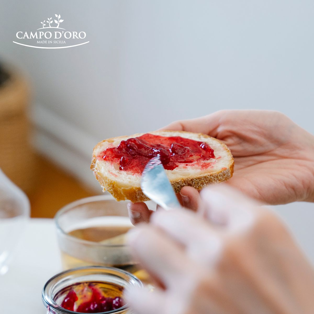 Person spreading red jelly on a slice of bread with 'Campo d'Oro' logo in the corner.