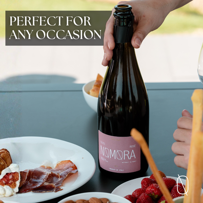 A hand holds a dark glass bottle of Nomora Rose Non-Alcoholic Sparkling Wine with a pink label, set on a dark table with plates of food in the foreground and a text overlay that reads 'PERFECT FOR ANY OCCASION'.