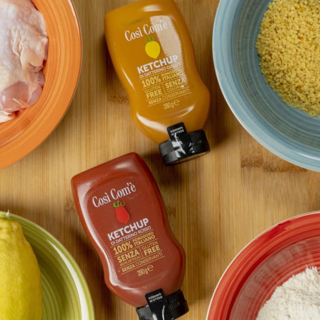 Two bottles of Così Com’è Italian Tomato Ketchup on a wooden surface — one made with red Datterino tomatoes (200 g) and the other with yellow Datterino tomatoes (180 g). Both are labeled gluten-free, preservative-free, and made with 100% Italian tomatoes.