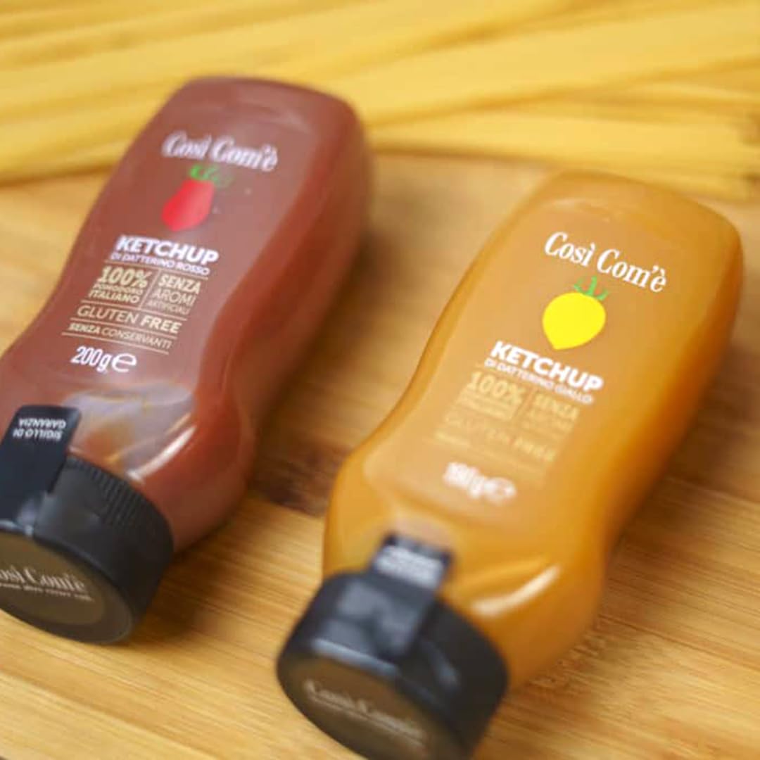 Two bottles of Così Com’è Italian Tomato Ketchup on a wooden surface — one made with red Datterino tomatoes (200 g) and the other with yellow Datterino tomatoes (180 g). Both are labeled gluten-free, preservative-free, and made with 100% Italian tomatoes.