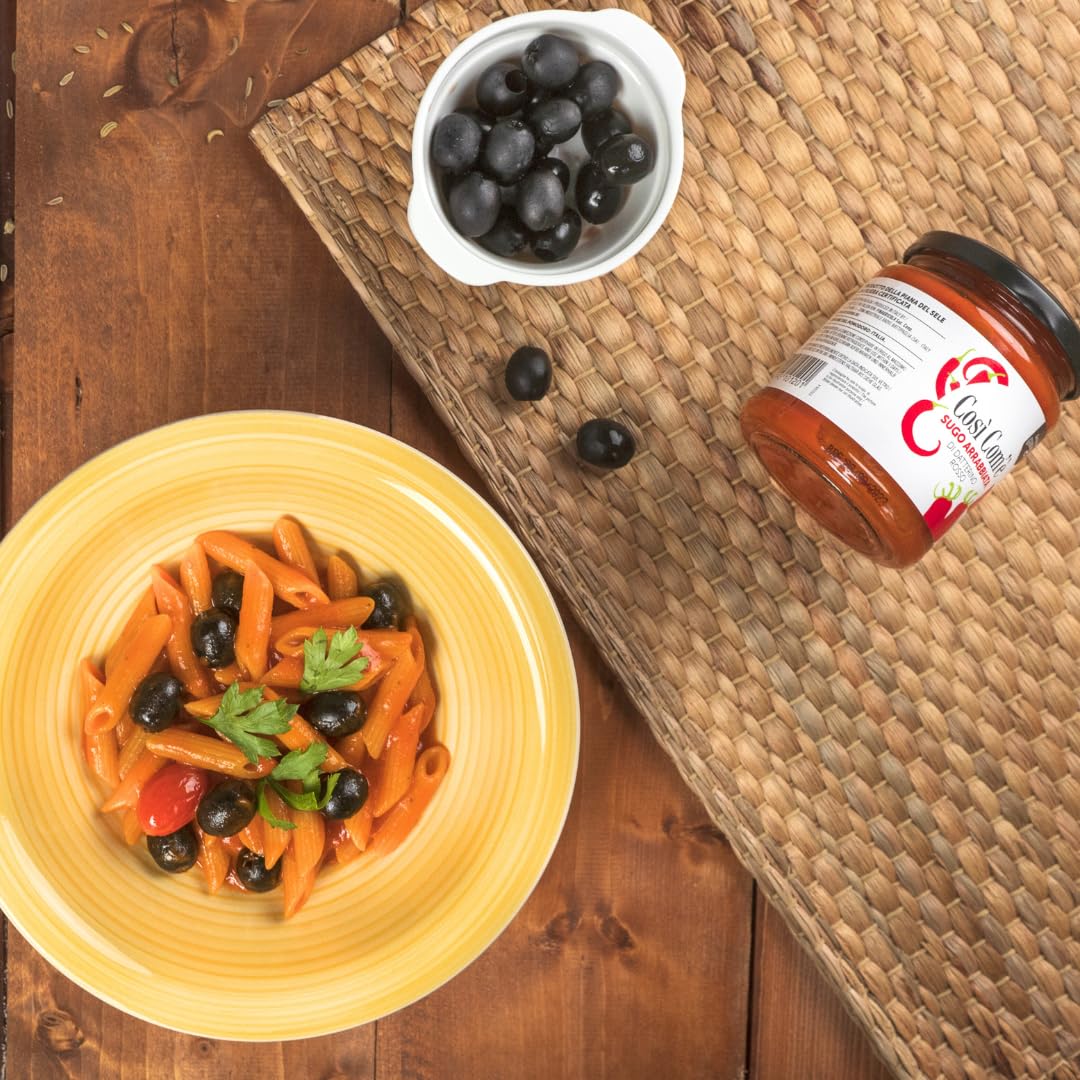 Yellow plate of penne pasta with black olives, cherry tomatoes, and parsley, next to a jar of Così Com’è Arrabbiata tomato sauce and a small bowl of fresh black olives on a wooden table.