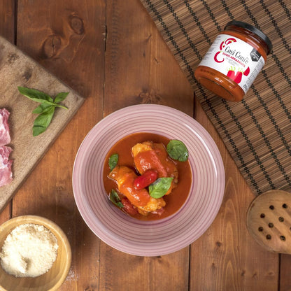 Così Com’è Arrabbiata red Datterino tomato sauce jar on a wooden table with a bowl of stuffed peppers in spicy tomato sauce, garnished with basil leaves, next to Parmesan cheese, fresh basil, and raw meat.