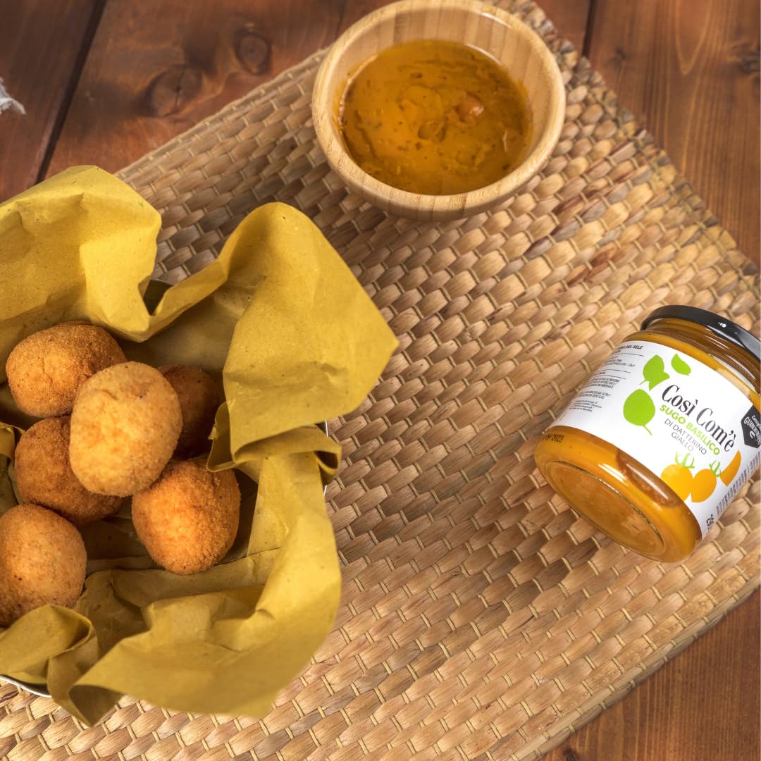 Così Com’è Yellow Datterino Tomato Sauce with Basil jar placed beside golden fried arancini and a wooden bowl of tomato sauce on a rustic woven mat