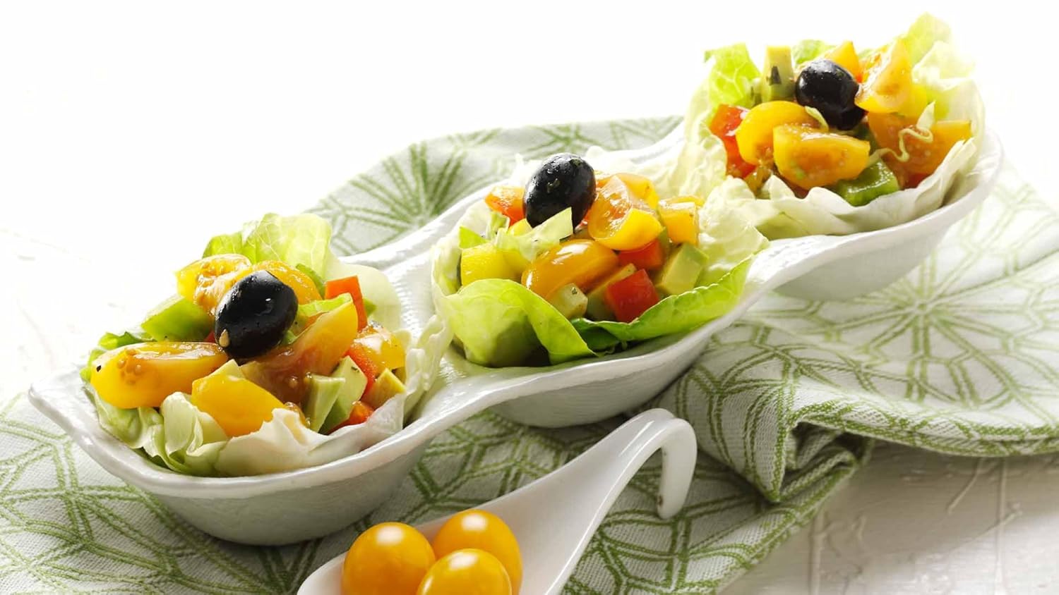 Fresh salad cups with yellow datterino tomatoes, avocado, bell peppers, black olives, and lettuce leaves, served in a white dish on a patterned green cloth