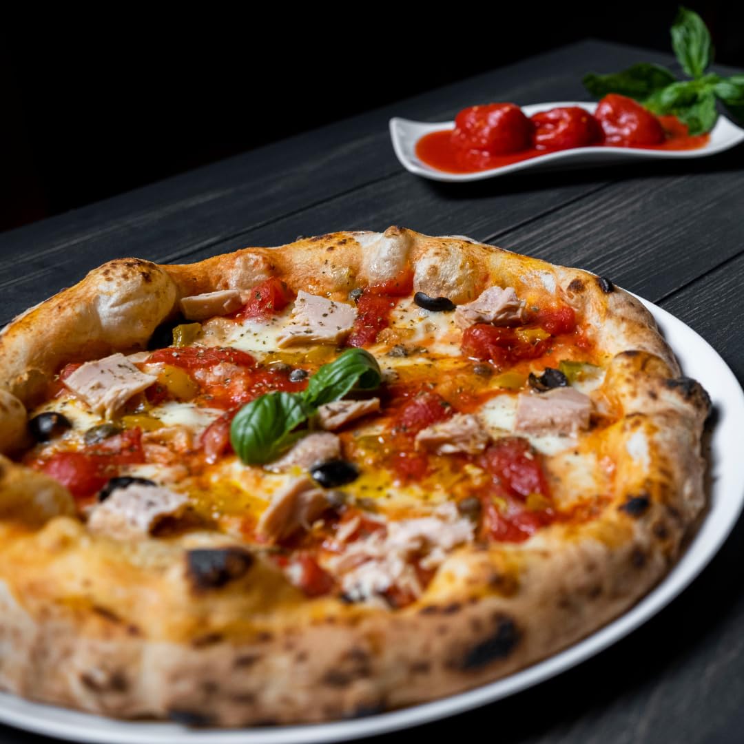 Close-up of an artisan pizza topped with red Datterino tomatoes, mozzarella, tuna chunks, black olives, and fresh basil leaves, served on a white plate with whole peeled tomatoes in sauce and fresh basil in the background.