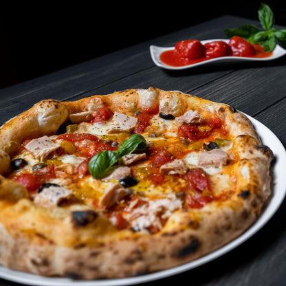 Close-up of an artisan pizza topped with red Datterino tomatoes, mozzarella, tuna chunks, black olives, and fresh basil leaves, served on a white plate with whole peeled tomatoes in sauce and fresh basil in the background.