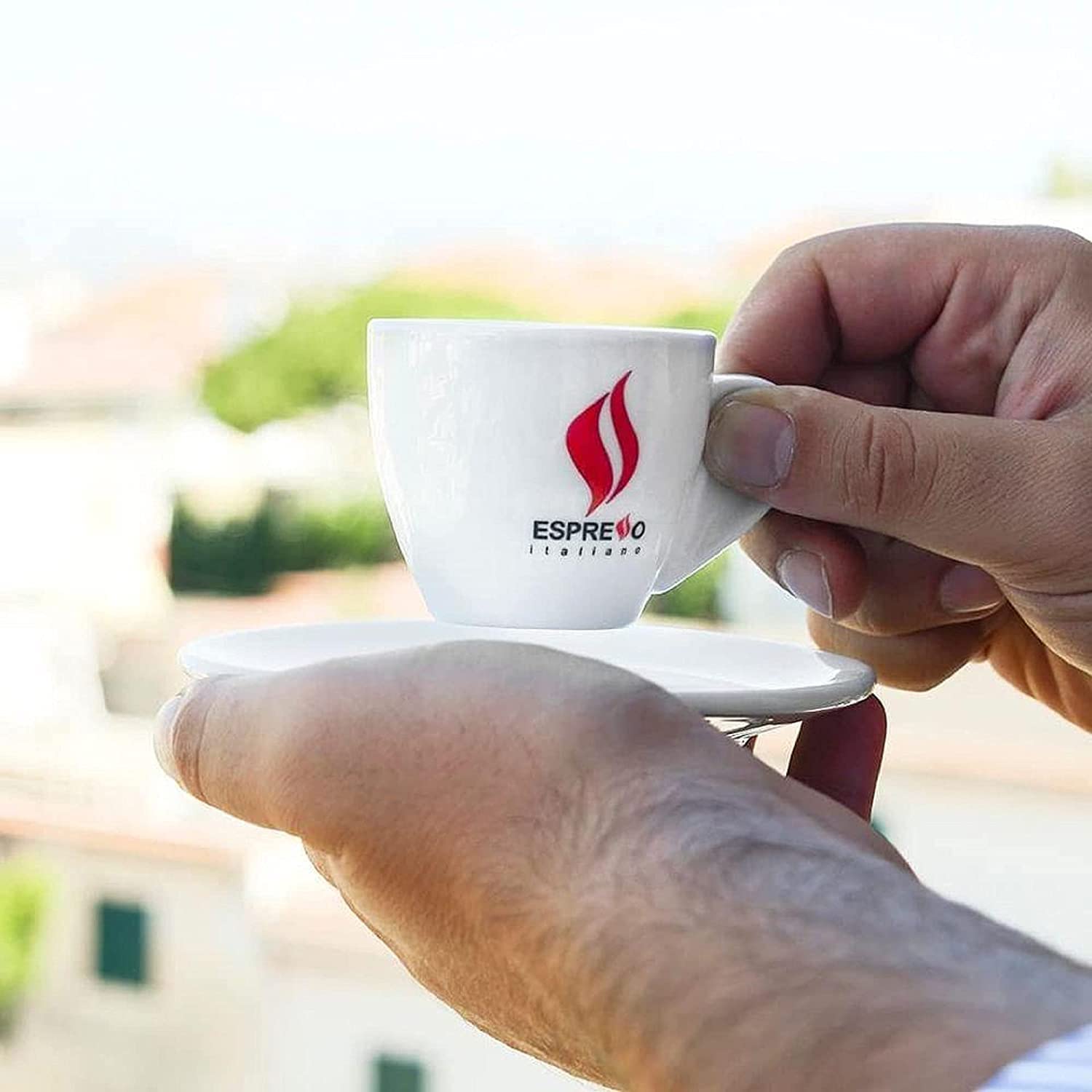Close-up of a person holding a white espresso cup and saucer with the Espresso Italiano logo, enjoying authentic Italian coffee outdoors.