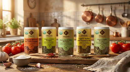 Row of organic spice containers on a wooden board with tomatoes and kitchen utensils in the background.