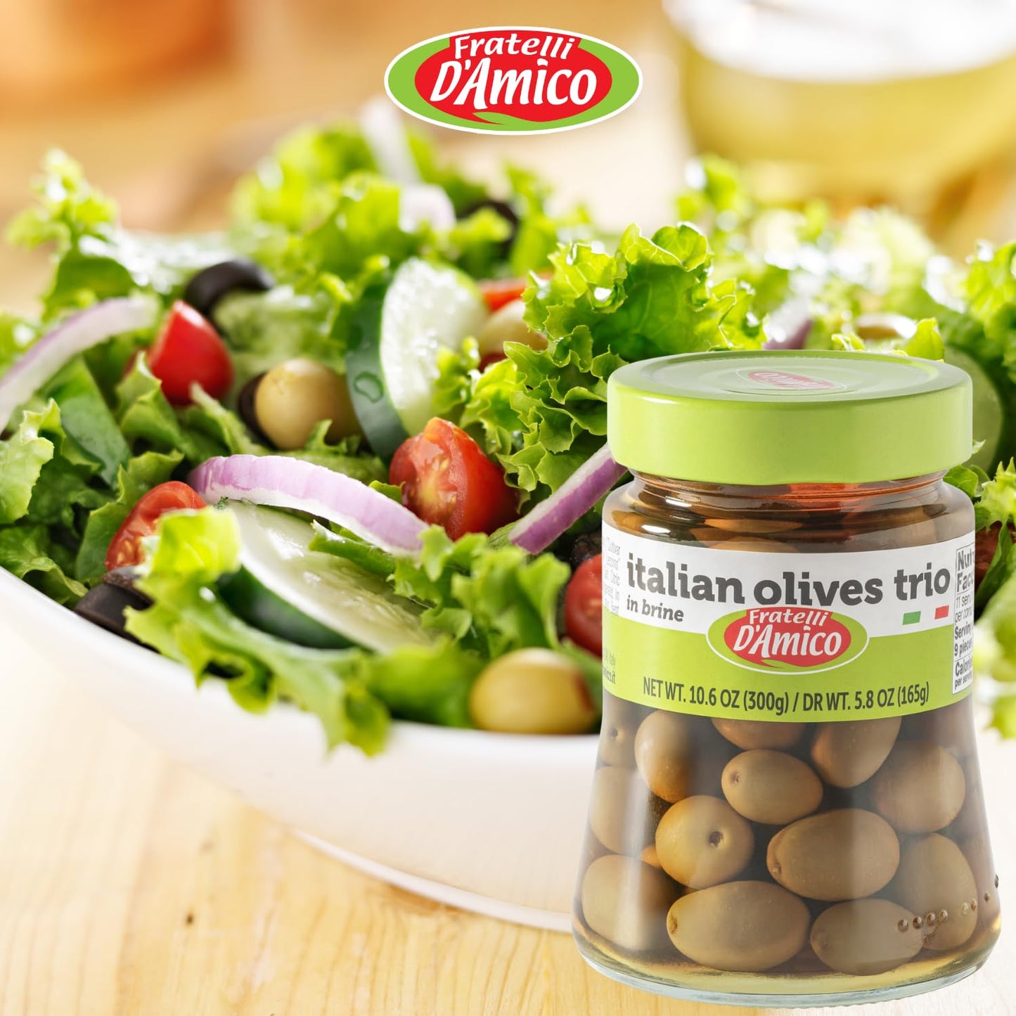 A fresh Mediterranean salad with lettuce, cherry tomatoes, cucumber slices, red onion, and black olives is served in a white bowl. In the foreground, a glass jar of Fratelli D’Amico Italian Olives Trio in brine (10.6 oz / 300g) is displayed, emphasizing its authentic Italian origin and quality. The Fratelli D’Amico logo appears at the top of the image.