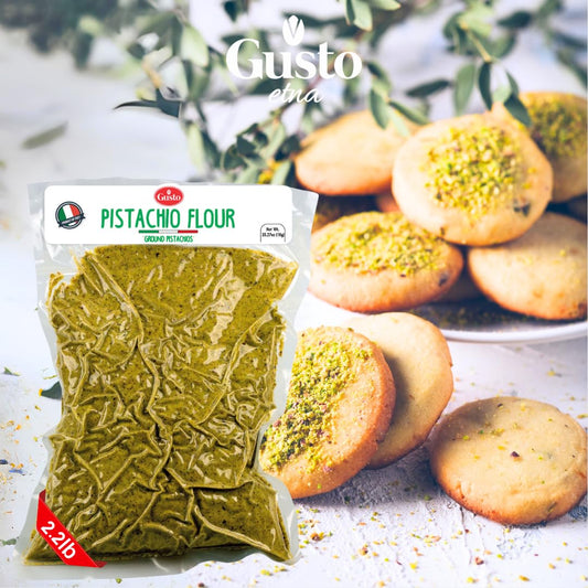 Clear plastic bag containing 2.2 lb (1 kg) of finely ground pistachio flour from the brand Gusto Etna, displayed in front of a plate of round cookies topped with crushed pistachios, with green leaves in the background