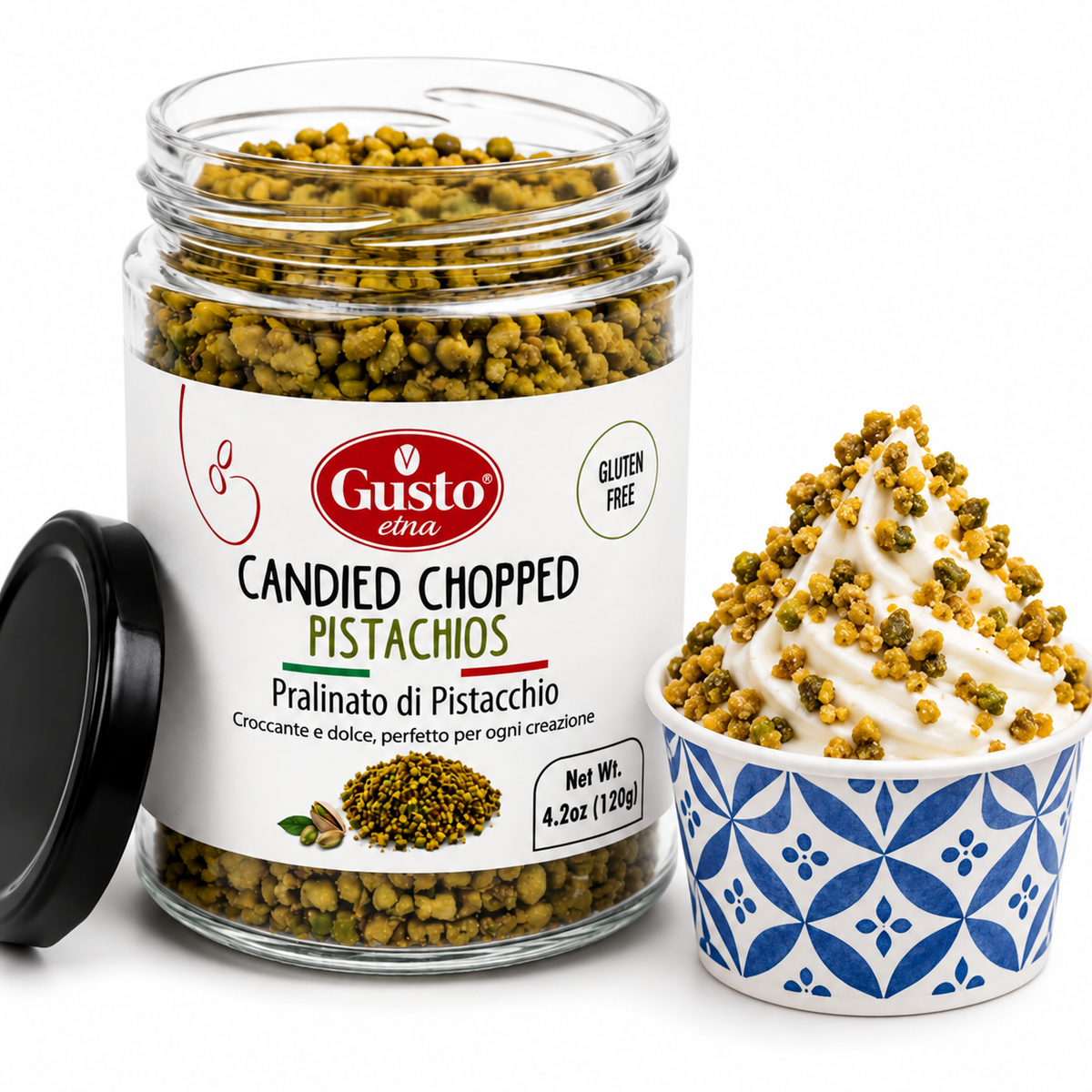 A glass jar of Gusto Eina Candied Chopped Pistachios with its black lid leaning beside it, next to a blue and white patterned cup filled with whipped cream topped with the candied pistachio pieces on a white background.