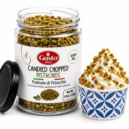 A glass jar of Gusto Eina Candied Chopped Pistachios with its black lid leaning beside it, next to a blue and white patterned cup filled with whipped cream topped with the candied pistachio pieces on a white background.