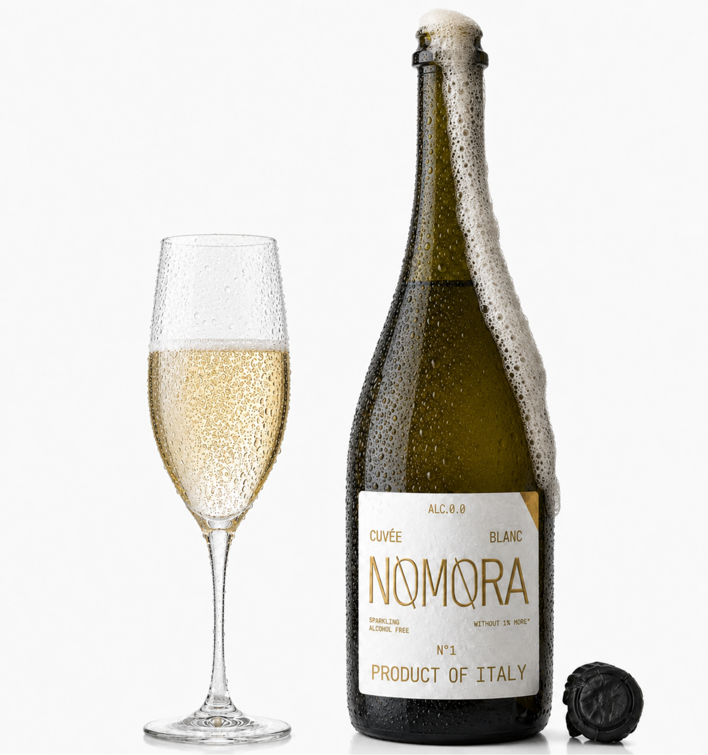 A bottle of Nomora sparkling wine with a foaming neck and a filled flute glass beside it on a clean white background. The bottle has a dark green glass body, a white label with gold lettering, and a black cap lying nearby.