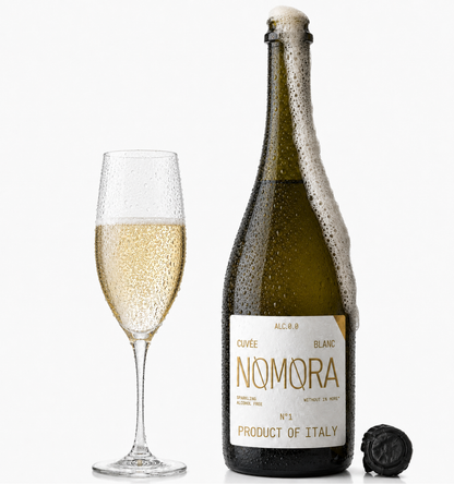 A bottle of Nomora sparkling wine with a foaming neck and a filled flute glass beside it on a clean white background. The bottle has a dark green glass body, a white label with gold lettering, and a black cap lying nearby.
