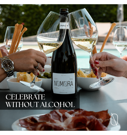 A dark wine bottle labeled NOMORA stands on a table with two people's hands holding wine glasses filled with white wine, surrounded by breadsticks, olives, crackers, and sliced cured meat. Text on the image reads 'CELEBRATE WITHOUT ALCOHOL'.