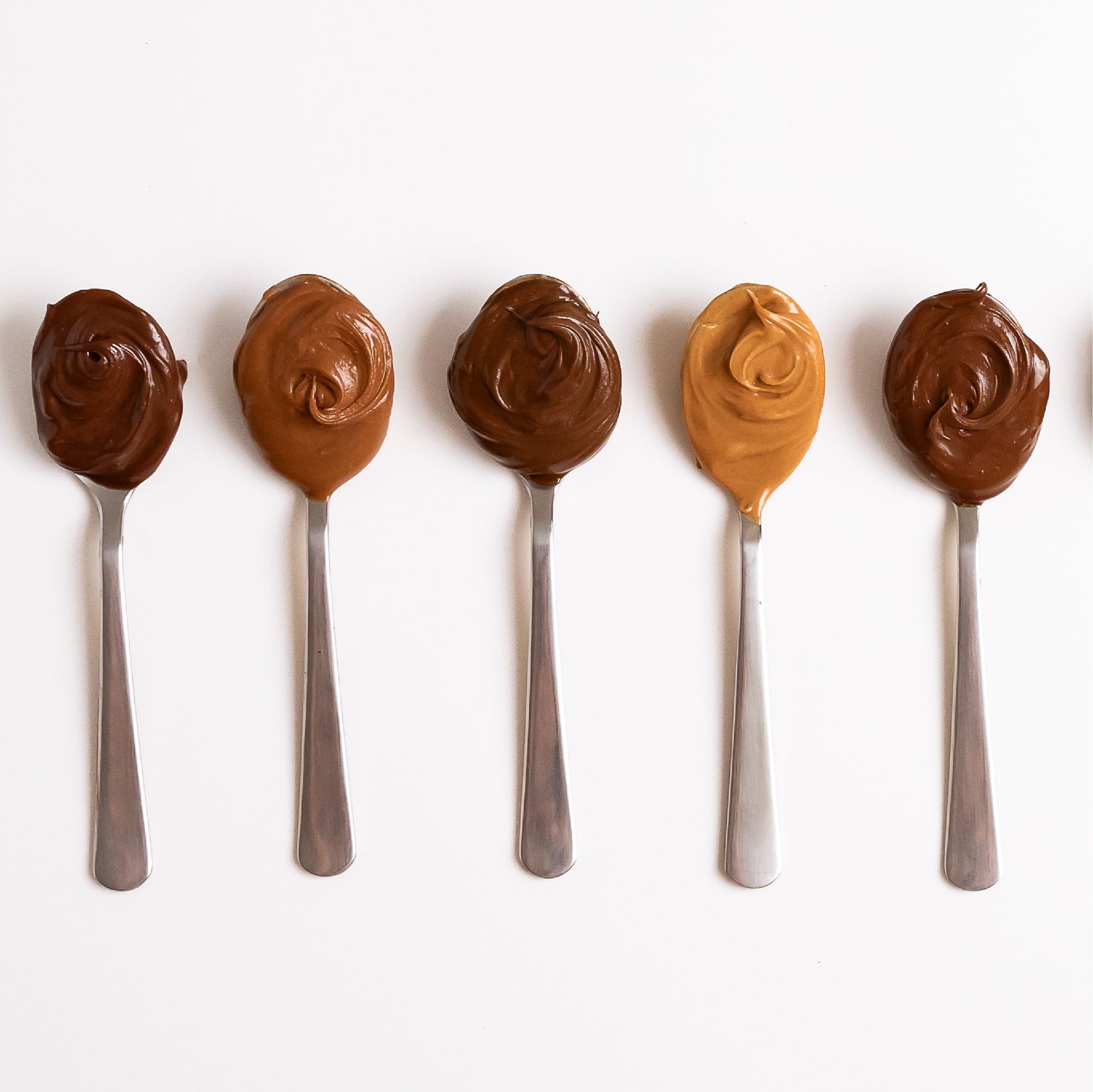 Five metal spoons lined up on a white background, each filled with different shades of Nutlicious hazelnut spreads, showcasing a variety of flavors and textures.