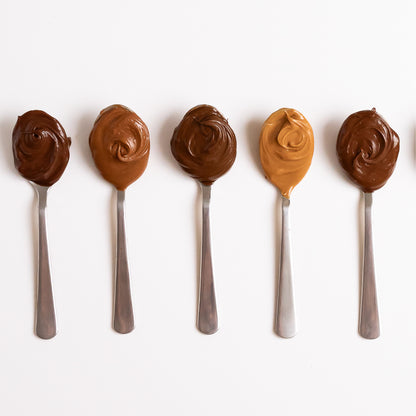 Five metal spoons lined up on a white background, each filled with different shades of Nutlicious hazelnut spreads, showcasing a variety of flavors and textures.