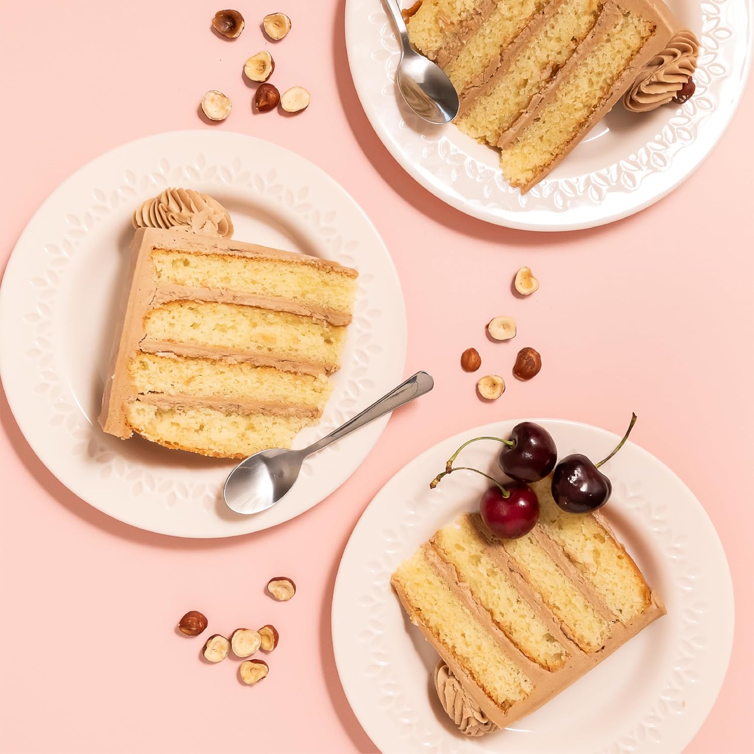 Plates with slices of hazelnut-flavored layer cake filled and frosted with Nutlicious spread, decorated with cherries and hazelnuts on a pink background.