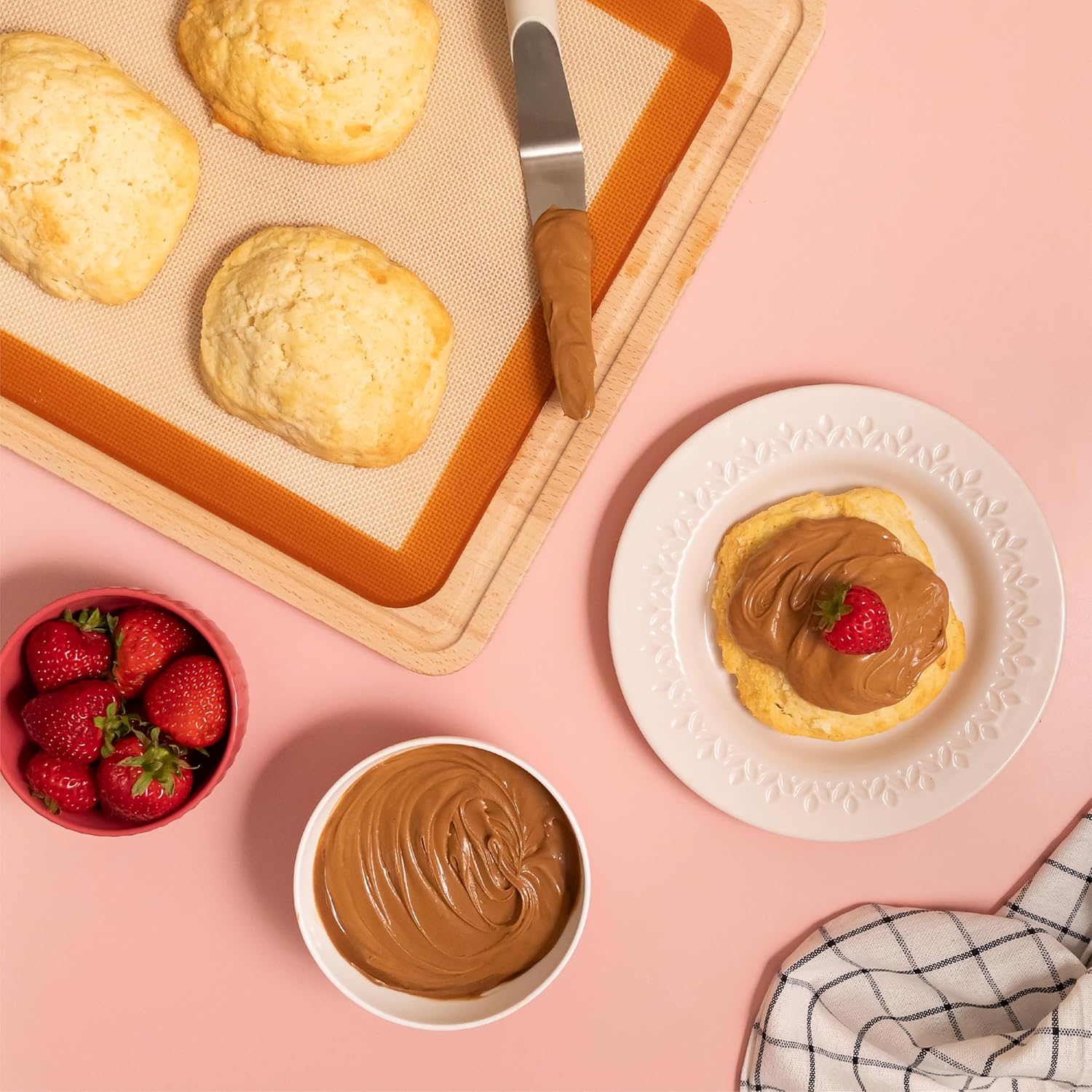 Homemade biscuits served with creamy Nutlicious hazelnut spread and topped with fresh strawberries, surrounded by a bowl of spread and whole strawberries on a pink background.