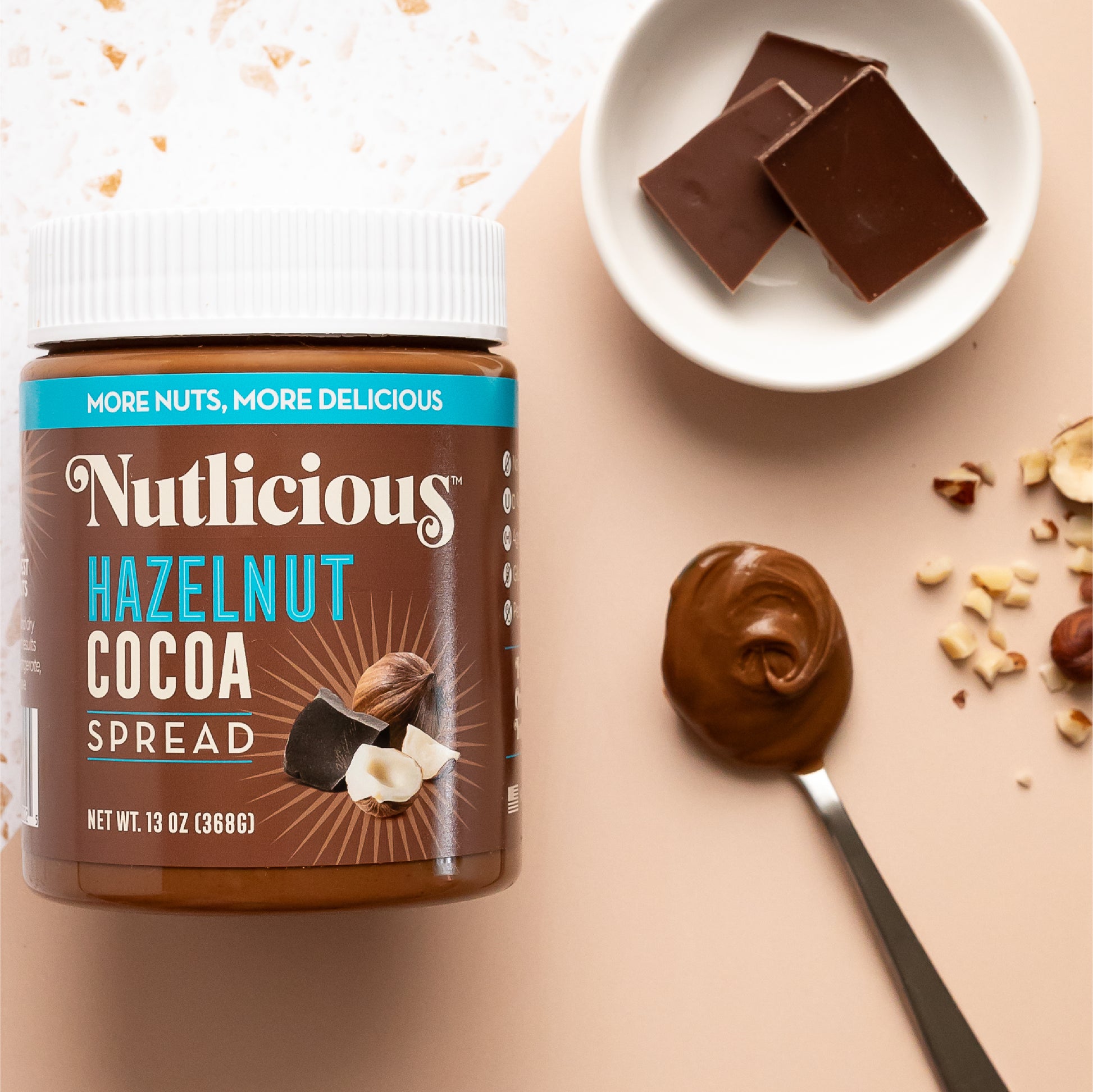 Nutlicious Hazelnut Cocoa Spread (13 oz) jar displayed on a flat surface with a spoonful of creamy spread, chocolate squares in a small dish, and scattered chopped hazelnuts for decoration.