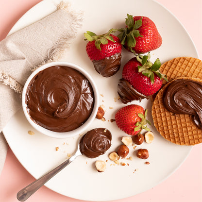 Plate with fresh strawberries dipped in hazelnut dark cocoa spread, waffle cookies topped with spread, a bowl of chocolate hazelnut spread, and scattered hazelnuts, creating a delicious dessert scene.