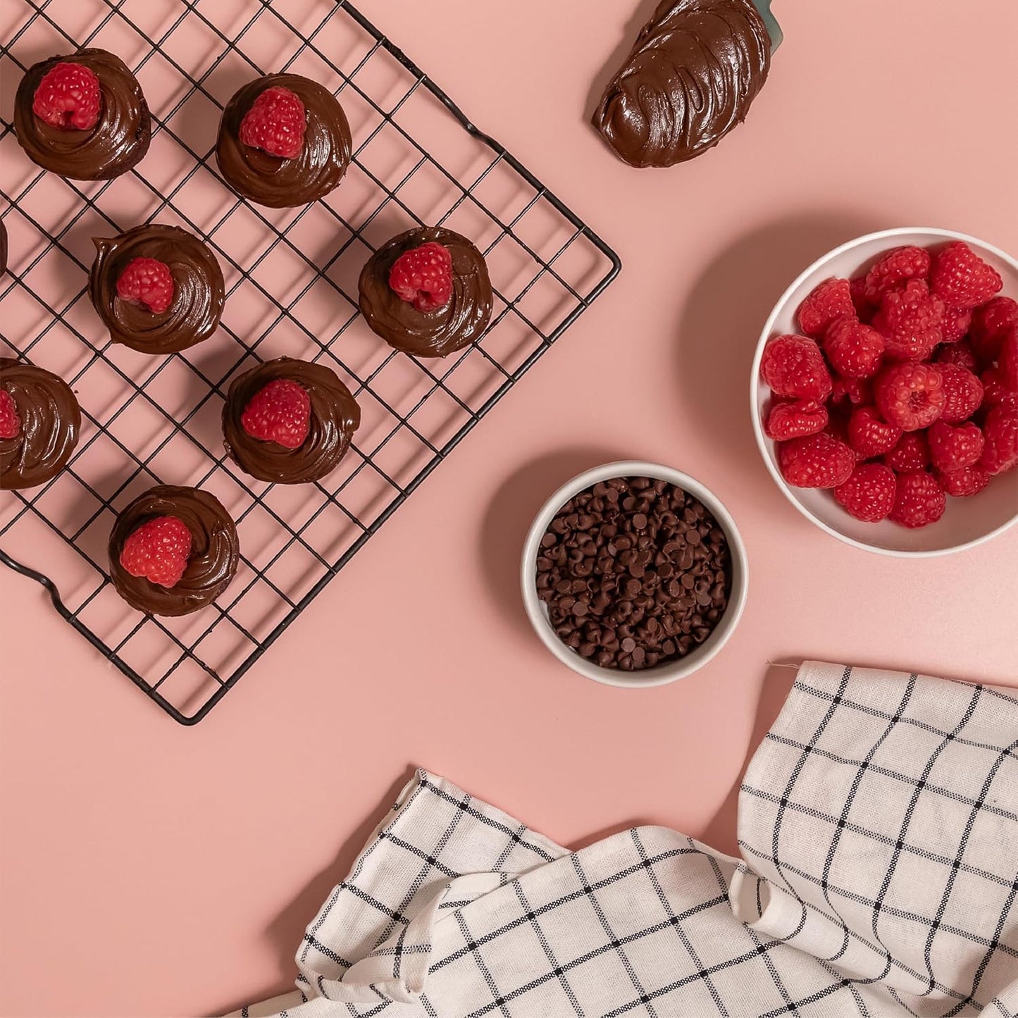 Cupcakes topped with hazelnut espresso spread and raspberries on cooling rack, with bowls of mini chocolate chips and raspberries on pink background.